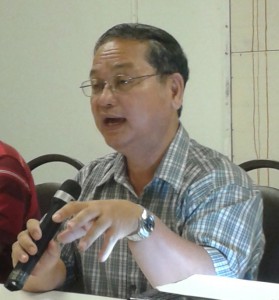 A person wearing glasses and a plaid shirt is seated at a table, passionately speaking into a microphone. With one hand gesturing, papers sprawled before them, they discuss the vital role of indigenous communities in preserving Borneo's lush rainforest.